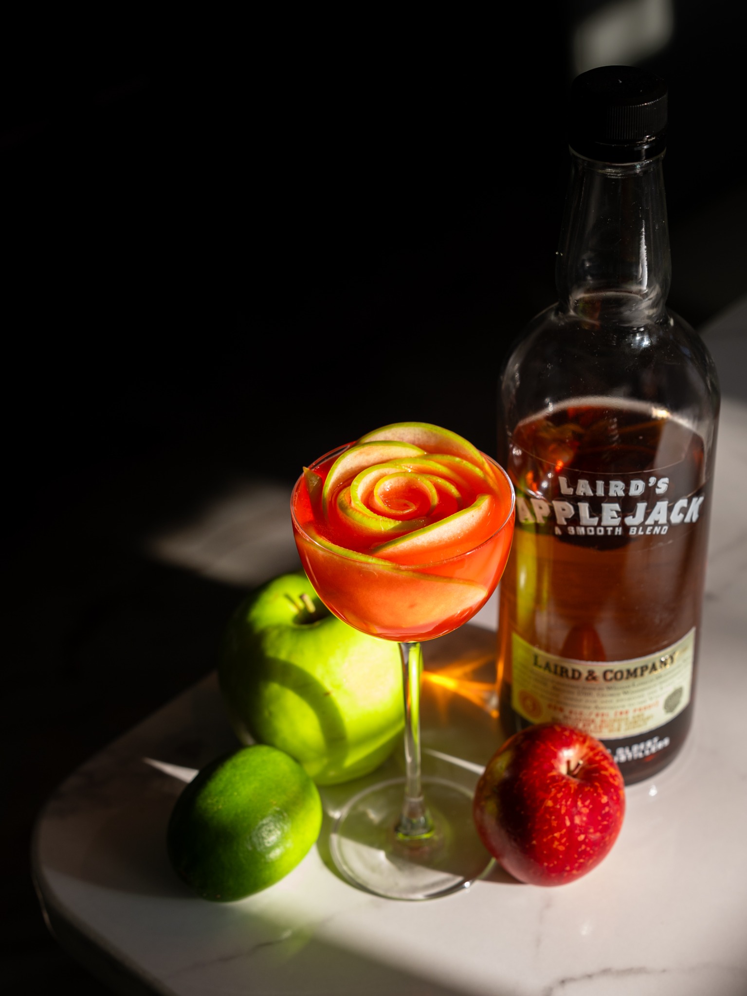 Orange cocktail with a rose-shaped garnish beside an applejack bottle